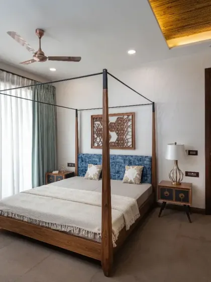 The guest bedroom in Ashraya Residence is simple yet elegant. It features a wooden poster bed, a floral couch, and customized metal artwork, with rustic stone flooring lending a clean, grounded feel.