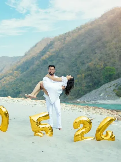 A classic pre-wedding shot on a river beach, with the groom lifting his bride in front of large foil balloons displaying their wedding date.