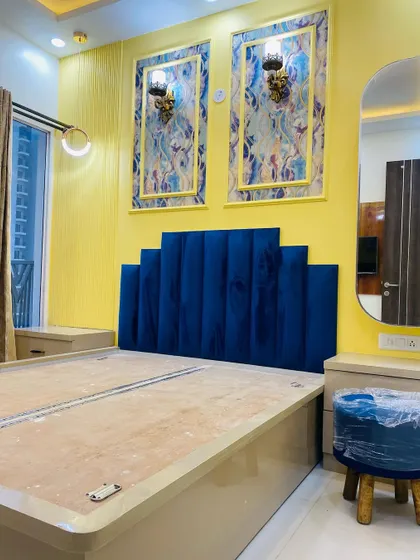 A bold and beautiful master bedroom design. The royal blue velvet headboard stands out against the vibrant yellow wall, which is decorated with framed panels and classic wall lights.