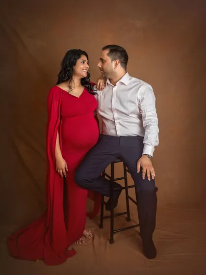 A classic and elegant studio maternity portrait of a couple. The woman, in a striking red gown, looks at her partner with a smile, creating a timeless and romantic image.