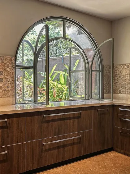 The kitchen features a large arched window above the counter, bringing in natural light and a view of the garden. The design combines wooden cabinetry with a patterned tile backsplash for a warm, classic feel.