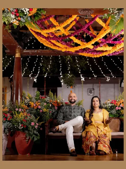 A magical Mehendi night comes to life with vibrant marigold garlands crisscrossing the ceiling, dotted with fairy lights. The seating area is surrounded by lush, colorful floral arrangements, creating a warm and festive atmosphere.