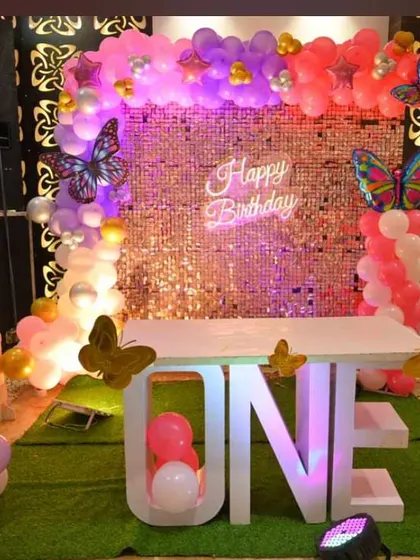 A first birthday setup with a butterfly theme on a green turf base. The decor features a "ONE" table, a pink sequin backdrop, and colorful butterfly accents.