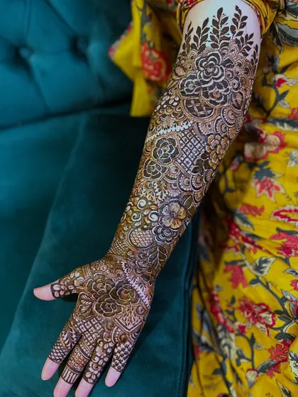 A full bridal henna design with floral motifs covering the entire arm. The yellow dress provides a vibrant backdrop for the dark henna.