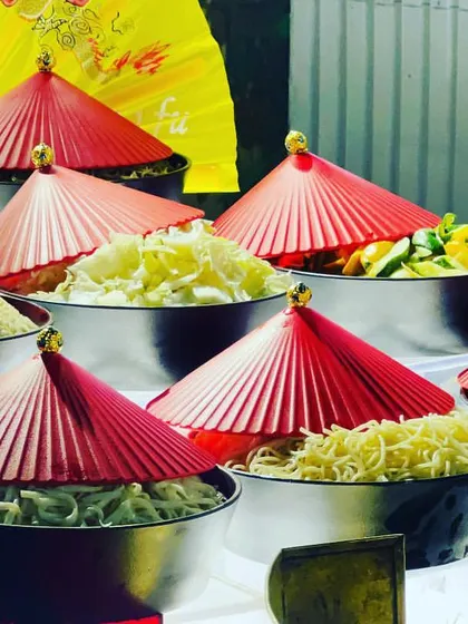 This live station features a variety of ingredients for a build-your-own Chinese bowl. The setup is adorned with traditional red paper hats and fans, adding a touch of authenticity and visual flair to the experience.