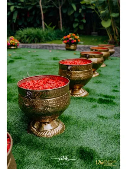 The aisle leading to the Haldi setup is lined with brass pots filled with red petals, adding a pop of color to the green lawn.