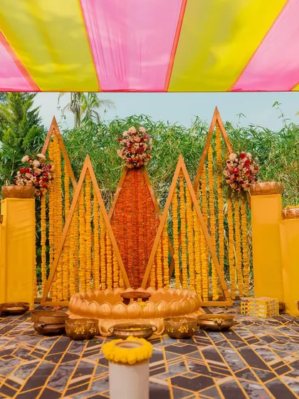 A geometric and colorful Haldi stage. We used triangular wooden frames decorated with marigold strings to create a modern yet traditional backdrop, complemented by brass bowls and a lotus seat for the bride or groom.