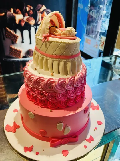 A three-tier pink baby shower cake with a quilted bassinet topper holding a sleeping baby.
