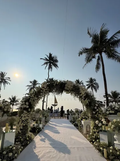 A beautiful floral archway frames the sea and sky at a beach wedding. The path is lined with white arrangements, creating a picturesque entrance to the ceremony.