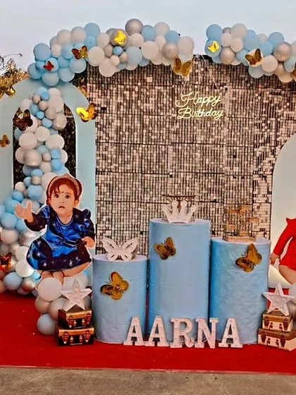 A beautiful butterfly theme for Aarna's first birthday. This outdoor setup features large butterfly wing cutouts, a silver sequin backdrop, and a blue and silver balloon garland, creating a stunning photo opportunity.