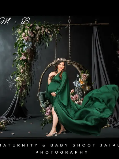 An elegant studio portrait featuring a woman in a flowing green gown on a floral-adorned swing. The dark, moody background creates a sophisticated and artistic atmosphere.