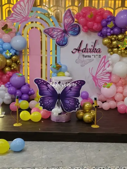A full view of the butterfly themed stage decoration for Advika's first birthday. The setup features a large backdrop, colorful balloon arrangements, and a light-up number '1'.