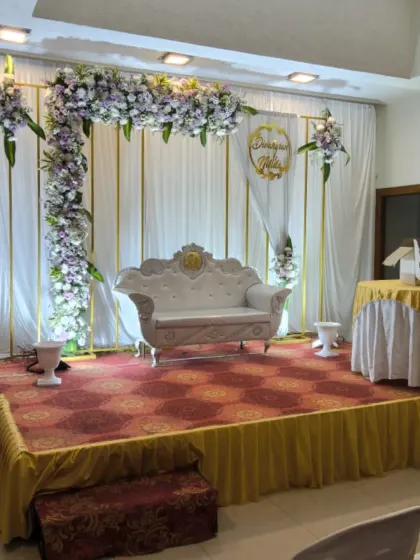 An elegant anniversary stage setup with a white and lavender floral frame, a classic white diwan, and simple white drapery.