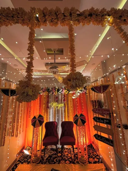 An intimate and traditional Haldi decor setup inside a home. The seating area is framed by orange drapes, marigold garlands, and fairy lights, with Rajasthani umbrellas adding a pop of color.