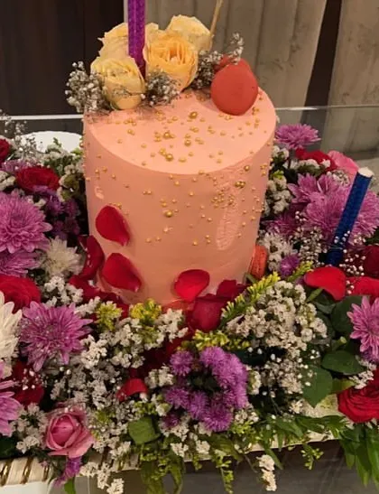 Gifting in style with this birthday cake platter. A tall pink cake is surrounded by a lush arrangement of roses, chrysanthemums, and baby's breath, creating a stunning and celebratory centerpiece.