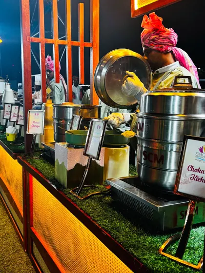 A live station serving traditional Gujarati dishes like Chokha nu Kichu. The setup uses a mix of modern steel warmers and traditional earthenware, set against a rustic, illuminated counter.