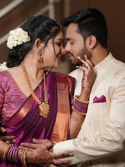 A beautiful, intimate moment between an engaged couple. Her makeup is classic and elegant, with a focus on defined eyes and a glowing complexion that complements her purple silk saree.
