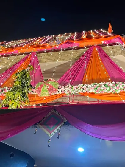 A house decorated with vibrant orange and pink drapes and fairy lights for a wedding. The decor also features colorful Rajasthani umbrellas.