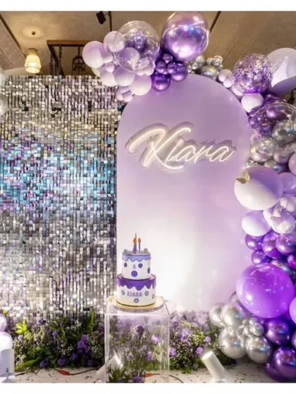 A personalized setup featuring a custom neon name sign. This backdrop combines a silver shimmer wall, a purple arch, and a balloon garland in shades of purple, silver, and white for a glamorous and unique look.