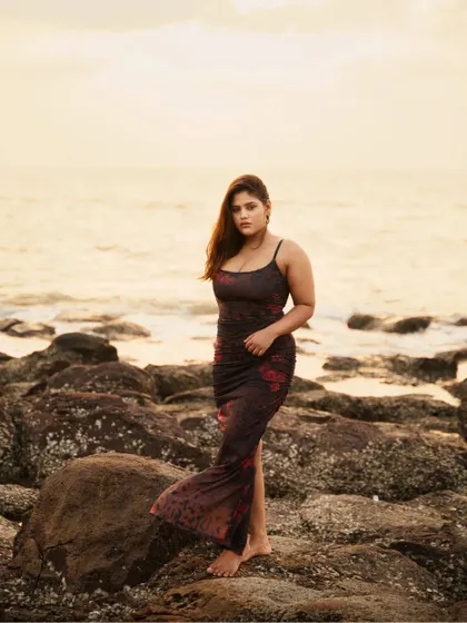 A full-body portrait on a rocky shoreline during sunset. The warm, golden light of the setting sun creates a beautiful, soft glow, highlighting the subject against the ocean backdrop.