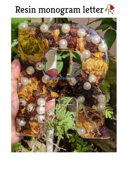 A resin monogram letter 'R' held up to the light, showing the beautiful transparency and the intricate details of the flowers preserved inside.