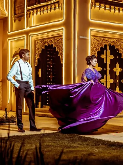 A dramatic, wide-angle shot of a couple during their pre-wedding shoot. The flowing purple metallic gown creates a stunning sense of movement against the illuminated architecture.