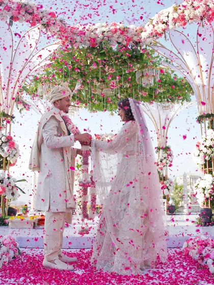 A perfect moment of joy as the couple exchanges garlands under a shower of pink petals. This image captures the essence of a dream wedding, with a stunning floral mandap as the centerpiece.