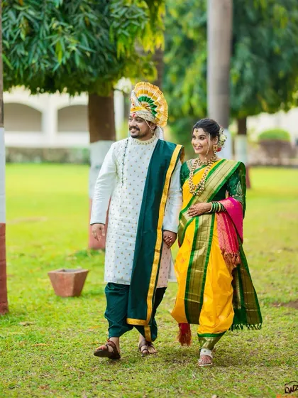 A candid shot of a Maharashtrian couple walking hand-in-hand through a garden, showcasing the groom's unique turban and their coordinated green and yellow outfits.
