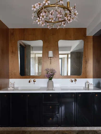 This bathroom is all about contrast, with a rich brown vanity and a black cabinet set against a veneered wood wall. The dual sinks and a delicate floral-inspired chandelier soften the space.