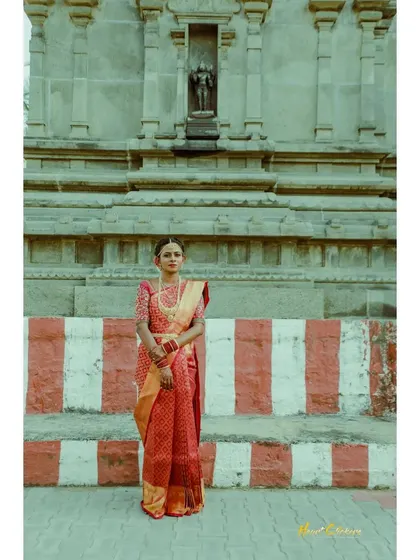 A solo portrait of the bride in her red silk saree, standing against the striking architecture of a temple.