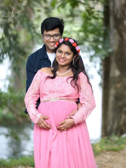A smiling couple posing by a lake. The mom-to-be is wearing a pink lace dress and a floral headband, and they both look so happy.