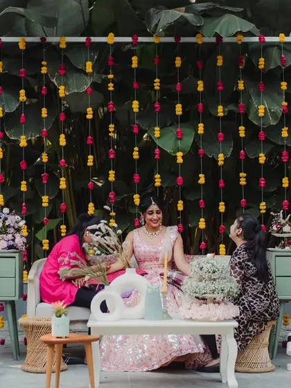 A chic and modern Mehendi setup with pastel furniture and a backdrop of colorful hanging flowers against lush greenery.