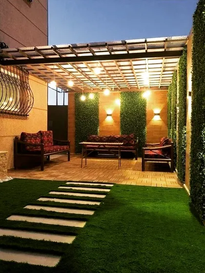 A narrow yard transformed with a pergola, artificial turf, and clever lighting. The back wall combines wood paneling and vertical gardens, illuminated by string lights and wall sconces to make the space feel larger.