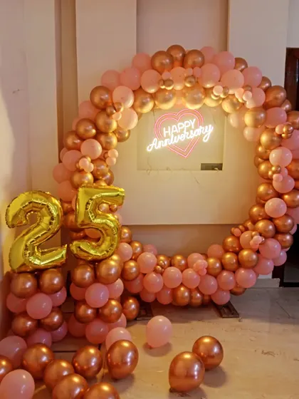 A beautiful 25th-anniversary decoration featuring a full ring of rose gold and pink balloons, a neon sign, and large '25' foil balloons.