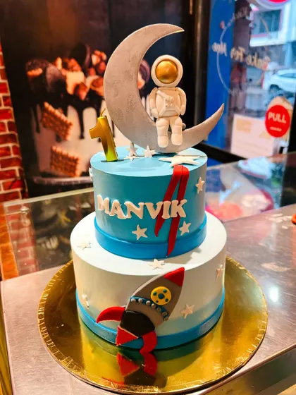 A two-tier astronaut cake for Manvik's first birthday, with a rocket ship and a cute astronaut sitting on the moon.