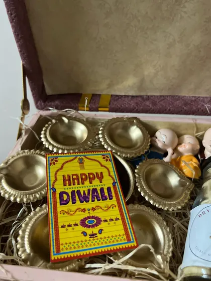 A close-up inside a pink velvet-lined box, filled with brass diyas, miniature monk figurines, and a 'Happy Diwali' card.
