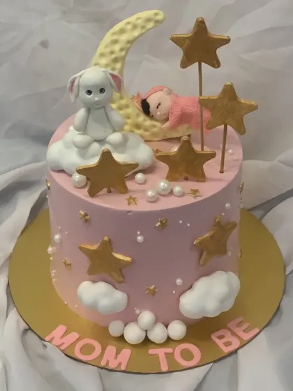 A lovely "Mom to Be" cake with a celestial theme. This pink cake features a sleeping baby on a moon, a bunny, and golden stars.