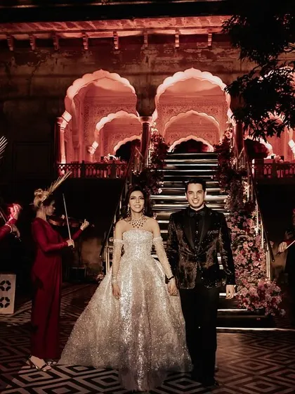The couple makes a royal entrance, flanked by a troupe of female violinists in dramatic red outfits, creating a powerful and elegant moment.