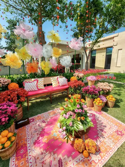 A stunning photo backdrop for the Mehendi, featuring a lush carpet of colorful flowers, a pink patterned rug, and a wall of giant, whimsical paper flowers in pastel shades.
