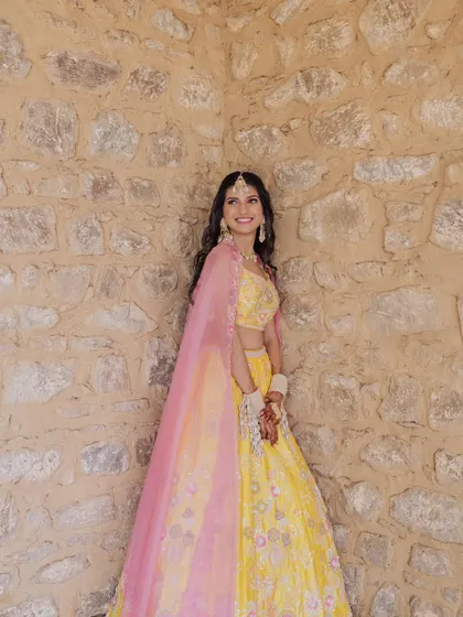 A lovely portrait of the bride in her yellow lehenga, her bright smile matching her vibrant outfit, captured against a rustic stone wall.