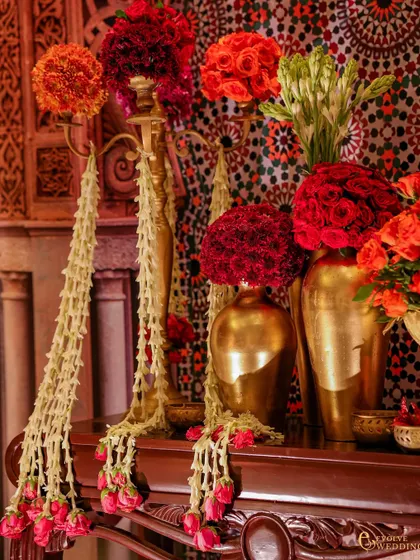 An ornate console table with cascading jasmine garlands and arrangements of red and orange flowers in golden vases. The mosaic backdrop adds a rich layer of pattern and color.