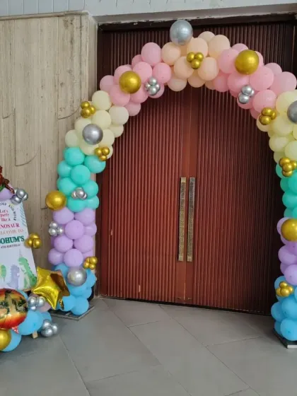 A pastel-colored balloon arch for a dinosaur-themed party entrance. The arch is decorated with gold and silver accents and placed next to a custom welcome board.