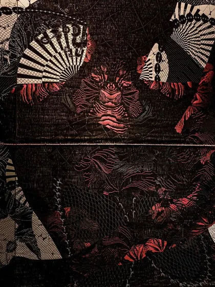 A detailed close-up of the 'Gothilicious' textile. The design, inspired by tattoo art, features intricate patterns in black, grey, and red, projecting a bold and glamorous persona.