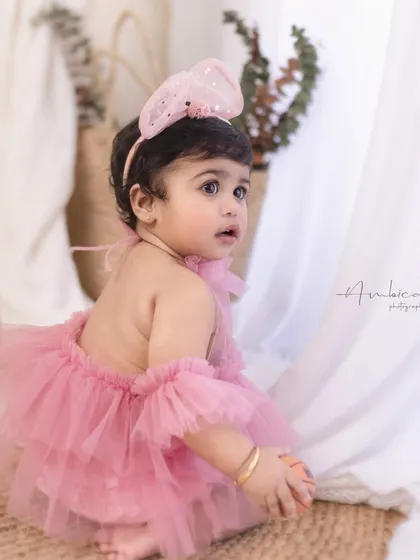 A baby girl looks over her shoulder in a beautiful pink romper. The soft side lighting highlights her features and the delicate texture of her outfit.