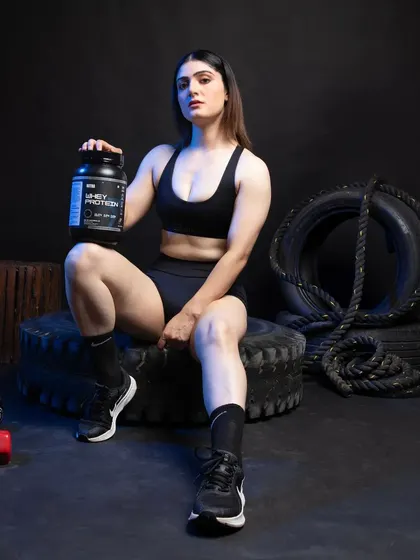 A female model sitting on gym tires, holding a whey protein container. The strong, direct pose is ideal for marketing materials that need to grab attention.