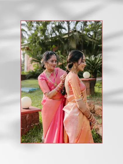 A touching moment between a bride and her mother, both in matching pink sarees, as her mother helps her get ready in a garden.
