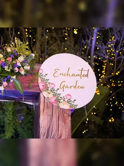 A whimsical detail from a birthday party, this "Enchanted Garden" sign sets the theme for the celebration. The sign is mounted on a wooden post with a small floral accent, surrounded by fairy lights.