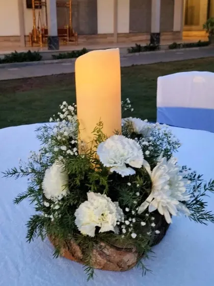 A simple centerpiece featuring a large pillar candle surrounded by white carnations and baby's breath, perfect for creating a warm and intimate atmosphere.