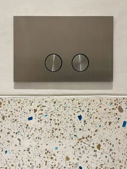 A minimalist flush plate is set against a colorful terrazzo wall. This juxtaposition of a sleek, modern fitting with a playful, textured material is a deliberate design choice to create visual interest.
