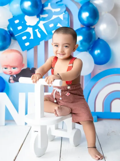 A happy little boy on a tricycle during his "Boss Baby" themed first birthday photoshoot. The blue and white balloon wall creates a festive backdrop.